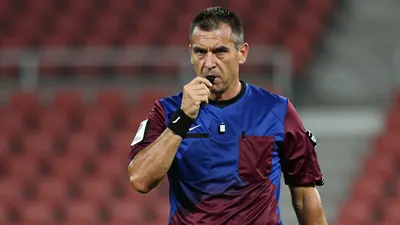 Middle-aged referee, muscular build, mid-30s, waist-up, wearing a plain training top split blue left side, claret right...