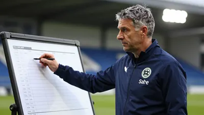 A mid-30s anonymous St Mirren coach in a dark blue training jacket, waist-up, focused on a tactical board, background...