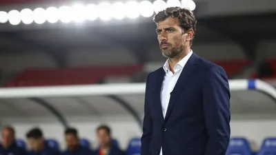 A 30-year-old male football manager in a navy blue suit with a white shirt, standing on the sidelines during a match...