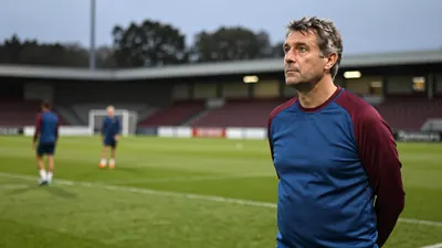 A 50-year-old male coach with a rugged build, wearing a plain blue and claret cotton training top, stands intensely on...